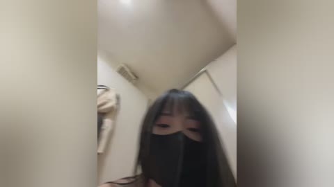A blurry video of a young woman with long black hair, wearing a black mask, standing in a dimly lit hallway. A beige jacket hangs on a hook to the left.