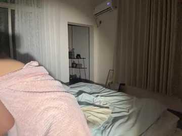 A video of a dimly lit bedroom with beige walls, an air conditioner, a partially open closet, and a person wearing a pink shirt and light gray sheets on the bed.