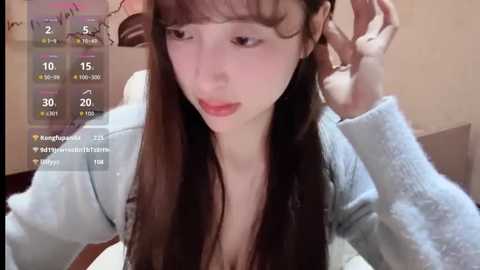 Video of a young, light-skinned Asian woman with straight, brown hair, wearing a light blue sweater, adjusting her hair. Background shows a beige wall and a white smartphone screen displaying social media metrics.