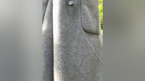 Video of a gray wool coat with a buttoned flap pocket, hanging in a blurred outdoor setting with green foliage in the background.