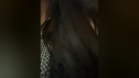 A dimly lit video of a person's neck and shoulder, with a patterned, dark fabric, possibly a checkered shirt. The background is blurred and dark, creating a moody atmosphere.