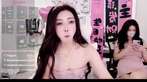 A video of an East Asian woman with long, dark hair and light skin, wearing a strapless pink dress and a tiara, sitting in a room with pink banners and a calendar.