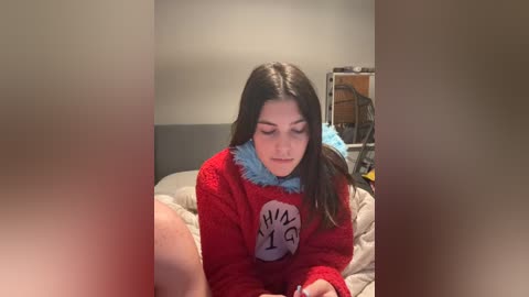 Video of a young woman with long dark hair wearing a red sweater with a blue collar, sitting on a bed in a dimly lit bedroom.