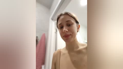 A video of a young woman with light skin and straight brown hair, smiling slightly. She is topless, standing in a modern bathroom with white walls and a pink towel hanging on the left.