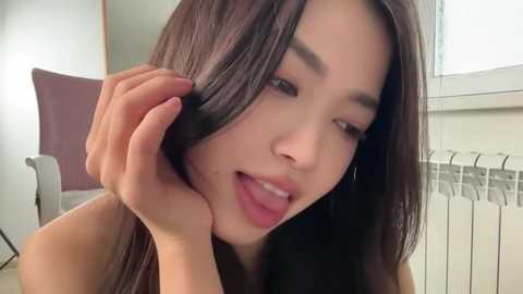 Video of a young Asian woman with long, straight black hair, wearing minimal makeup, playfully pulling her hair. Background shows a white chair and a window with white blinds.