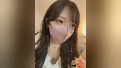 Video of an East Asian woman with long, straight black hair, wearing a pink surgical mask, white blouse, and necklace, indoors with a blurred background.