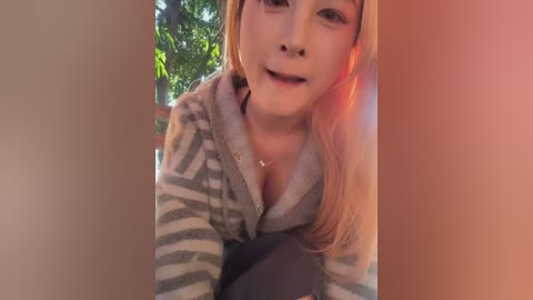 A video of a smiling young Asian woman with long blonde hair, wearing a grey striped hoodie, standing outdoors in sunlight with green foliage in the background.