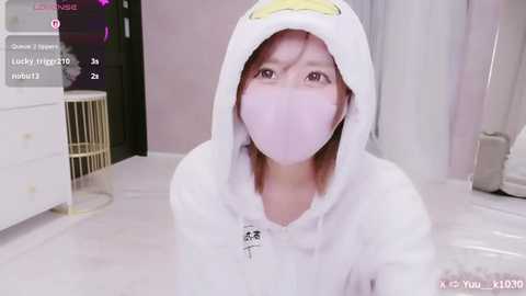 A video of a young woman with light skin, wearing a pink face mask and a white hoodie with a yellow duck design, sitting on a white floor in a room with a white dresser and a gold-framed mirror.