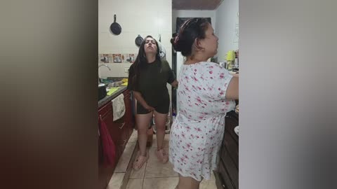 A video captures two women in a kitchen; one in a floral dress, the other in a dark outfit. The background features tiled walls, a sink, and cabinets.