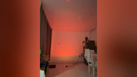 A video of a dimly lit, sparsely furnished bedroom with a red glow casting shadows on the white walls and floor. The room includes a bed, a chair, and scattered items, with a hint of clutter.