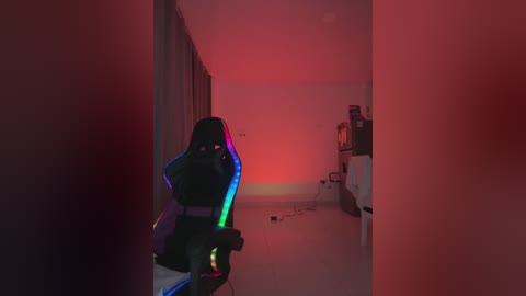 Video of a dimly lit, narrow hallway with a black gaming chair illuminated by a red glow, leading to a cluttered room with a computer desk.