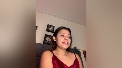 Video of a young woman with medium skin tone, dark hair, and a slender build, wearing a red satin camisole, standing indoors against beige walls with framed artwork.