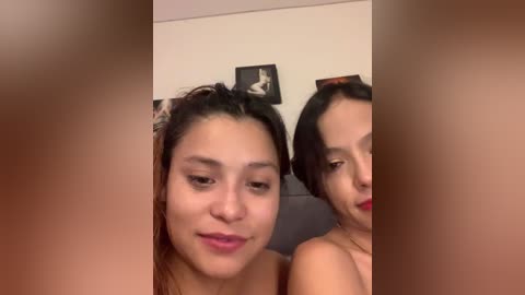 Video of two young women with light brown skin and dark hair, one smiling, the other looking away, in a dimly lit room with beige walls and a framed portrait.