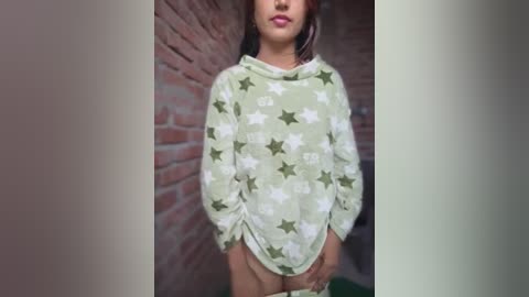 Video of a woman with medium skin tone and straight, dark hair, wearing a loose, star-patterned green and white sweater, standing in front of a brick wall, indoors.