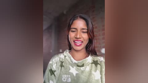 Video of a young woman with medium brown skin and straight dark hair, wearing a camouflage hoodie, smiling widely, set against a brick wall background.