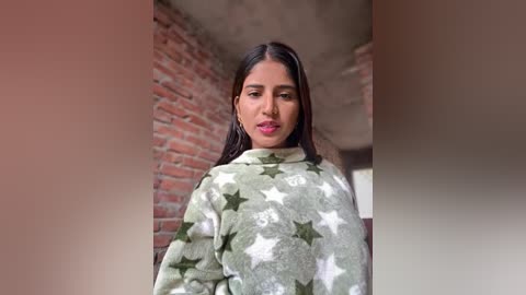 Video of a young woman with medium brown skin, long black hair, and full lips, wearing a camouflage-patterned hoodie, standing against a brick wall and concrete ceiling in a dimly lit room.