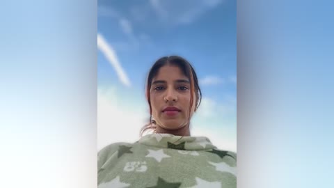 Video of a young Latina woman with medium brown skin and straight brown hair, wearing a green star-patterned hoodie, standing against a clear blue sky with a thin white contrail.