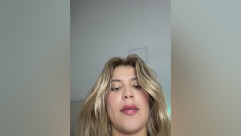 Video of a young woman with fair skin, wavy blonde hair, and a neutral expression, set against a plain, light-colored wall.