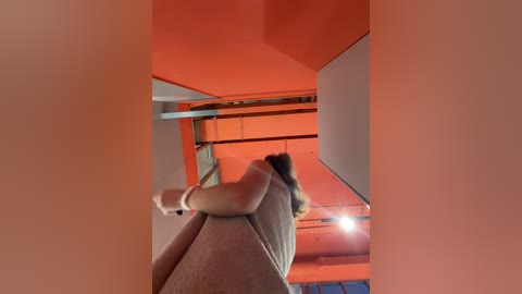 Media: Video of a woman with long hair in a gray tank top, sitting on a beige couch with her legs bound by a white rope, in a room with orange walls and ceiling.