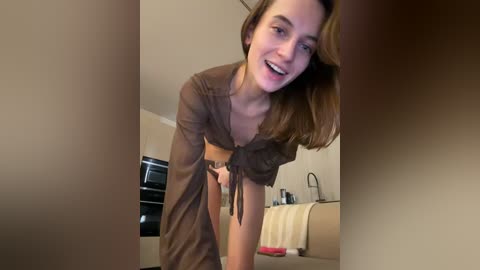 A video shows a smiling, light-skinned woman with brown hair, wearing a sheer, brown, open-front top and high-waisted shorts, leaning forward in a modern kitchen with beige walls and a black oven.