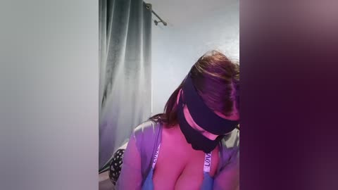 Video of a woman with brown hair, wearing a black blindfold, seated in a dimly lit room with grey curtains. She is dressed in a light grey satin robe.