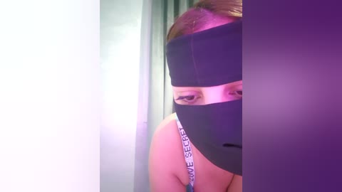 Video of a young woman with light skin and brown hair partially covered by a black blindfold. She wears a white tank top with \"VICTORIA'S SECRET\" written in black. The background is a dimly lit room with green curtains.