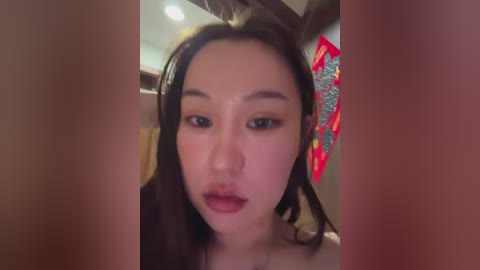 A close-up video of a young Asian woman with long, straight dark hair, light skin, and full lips, wearing minimal makeup, taken indoors with a blurred background.