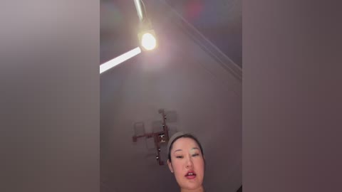 A video of a woman with East Asian features, light skin, and long dark hair, wearing makeup, standing in a dimly lit room with a ceiling fan, looking contemplative.