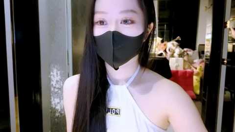 Video of an East Asian woman with long black hair, wearing a black face mask and a white sleeveless top with \"POPL\" text. She stands in a dimly lit room with a partially open door, revealing a colorful background with flowers and bags.