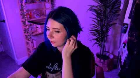 Video of a young woman with blue hair, wearing a black t-shirt, sitting in a dimly lit room with purple lighting, holding a phone to her ear, surrounded by plants and shelves.