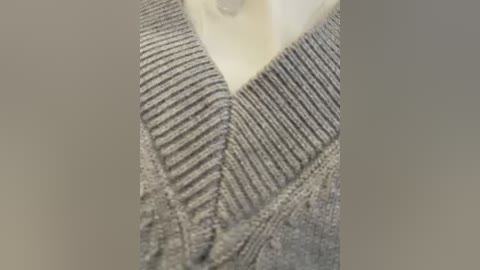 Video of a close-up view of a person's chest area, showcasing a V-neck, grey knitted sweater with textured, ribbed patterns, and a soft, cream-colored undershirt peeking out. The background is a smooth gradient of beige tones.