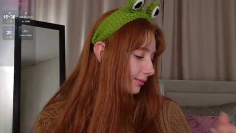 Video of a fair-skinned woman with long red hair, wearing a green frog hat, looking down at a phone, in a dimly lit room with beige curtains and a bed in the background.