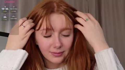 Video of a young woman with fair skin and long, reddish-brown hair, gently combing her hair with her hands, wearing a white sweater.