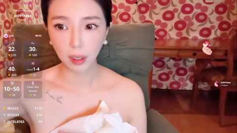 Video of a young East Asian woman with dark hair, wearing a white off-the-shoulder dress, sitting on a green chair, surrounded by retro floral-patterned wallpaper and a wooden table.