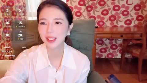 Video of an Asian woman with long black hair and light skin, smiling, wearing a white blouse, seated in a living room with retro red and white floral wallpaper, wooden desk, and green couch.