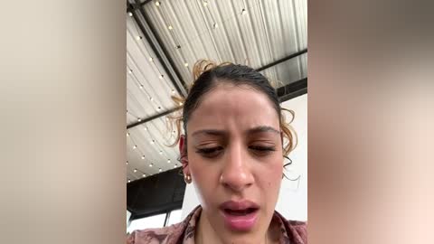 A video of a young woman with medium brown skin, curly hair, and hoop earrings, wearing a pink top, appears distressed with her mouth open, looking up at something. The background shows a corrugated metal roof and a black chair.