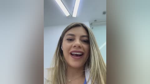 Video of a smiling, light-skinned woman with long, straight blonde hair, wearing a white lab coat, standing in a modern, well-lit room with fluorescent lights and white walls.