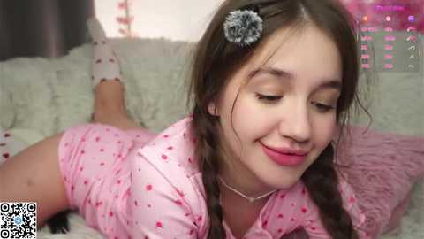Video of a young woman with fair skin, dark hair, and a flower headband, lying on her stomach in a pink polka-dot outfit, on a plush couch.