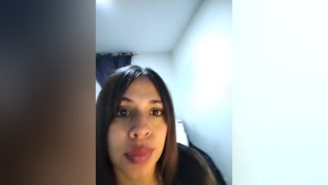 A close-up video of a young woman with light brown skin and long dark hair, making a pouty face, indoors with bright lighting and blurry background.