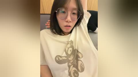 Video of an Asian woman with shoulder-length black hair, wearing large round glasses and a white T-shirt with a motorcycle graphic. She sits with one arm raised, revealing her side profile.