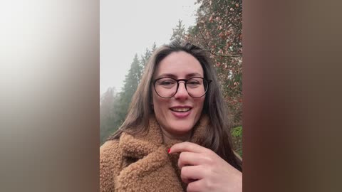 Video of a smiling woman with glasses, brown hair, and a tan jacket, standing in a forest with misty trees in the background.
