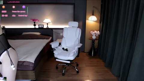 Video of a modern bedroom with a white leather gaming chair, a large bed, dark curtains, a lamp, and a TV screen displaying text.