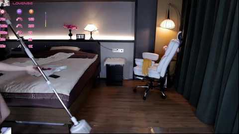 Video of a modern bedroom with a king-sized bed, white sheets, dark wooden floors, a white chair, and a black curtain, featuring a wall-mounted TV and dim lighting.