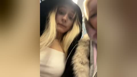 Video of a young blonde woman with long, straight hair, wearing a white off-the-shoulder top and black jacket, sitting on a bus, with blurred background and out-of-focus faces of other passengers.