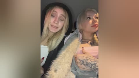 Video of two young women: blonde with a black hoodie, pale skin, closed eyes; blonde with tattoos, light skin, closed eyes, wearing a denim jacket, both smiling.