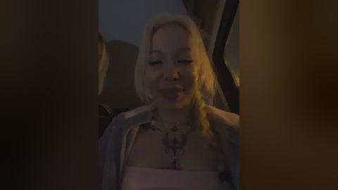 Video of a blonde woman with fair skin, wearing a low-cut top and a necklace, sitting in a dimly lit car, smiling softly.