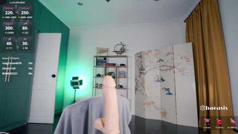 A video captures a modern, minimalist bedroom with a large, pale pink dildo on a bed, digital display screens showing statistics, and a white, floral screen divider.