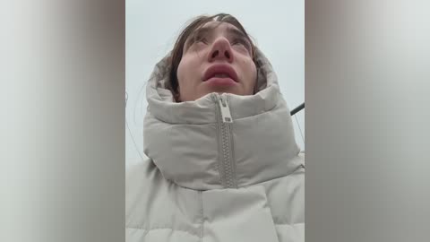 Video of a woman in a puffy, light beige winter coat, her face partially obscured by the hood. She has light skin and a serious expression, looking up with her head tilted back.