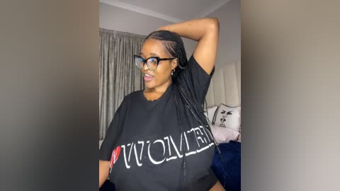 A video of a Black woman with medium-brown skin, wearing glasses, black T-shirt with \"WOMAN\" in white text, and long braids, standing in a bedroom with white walls and curtains.
