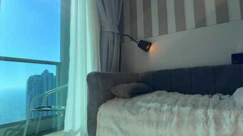 Video of a modern, minimalist bedroom with a bed covered in white and beige blankets, a gray upholstered headboard, and a lamp on the wall. Large windows with sheer curtains and a view of a city skyline in the background.